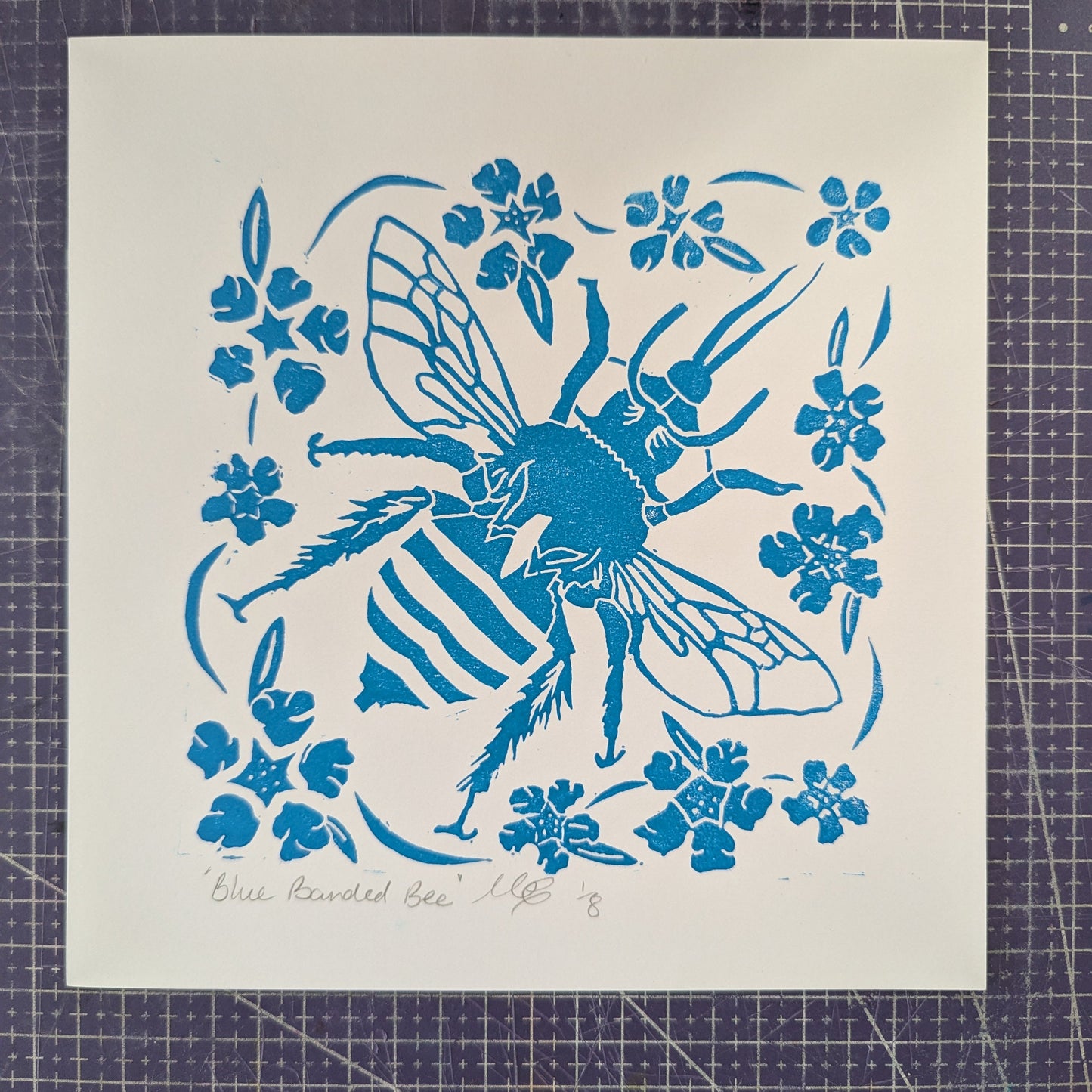 Blue bee and floral design on a white cardstock sheet, placed on a grid surface.